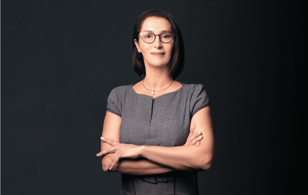 Professional headshot of Dr. Cynthia Poulos standing confidently with arms crossed, wearing glasses and a gray dress against a dark background.