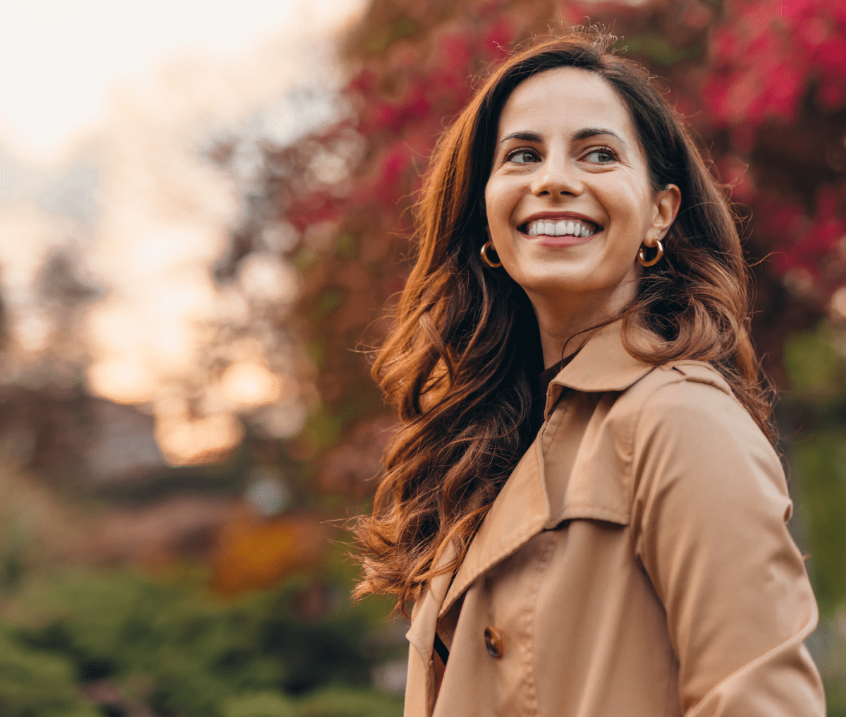 Smiling woman with long brown hair wearing a tan trench coat in an autumn setting.